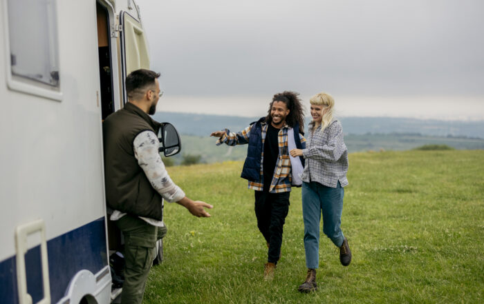 Happy couple walking across a meadow greeting a camper van owner who's waiting for them at the door of his vehicle, ready to start a road trip adventure
